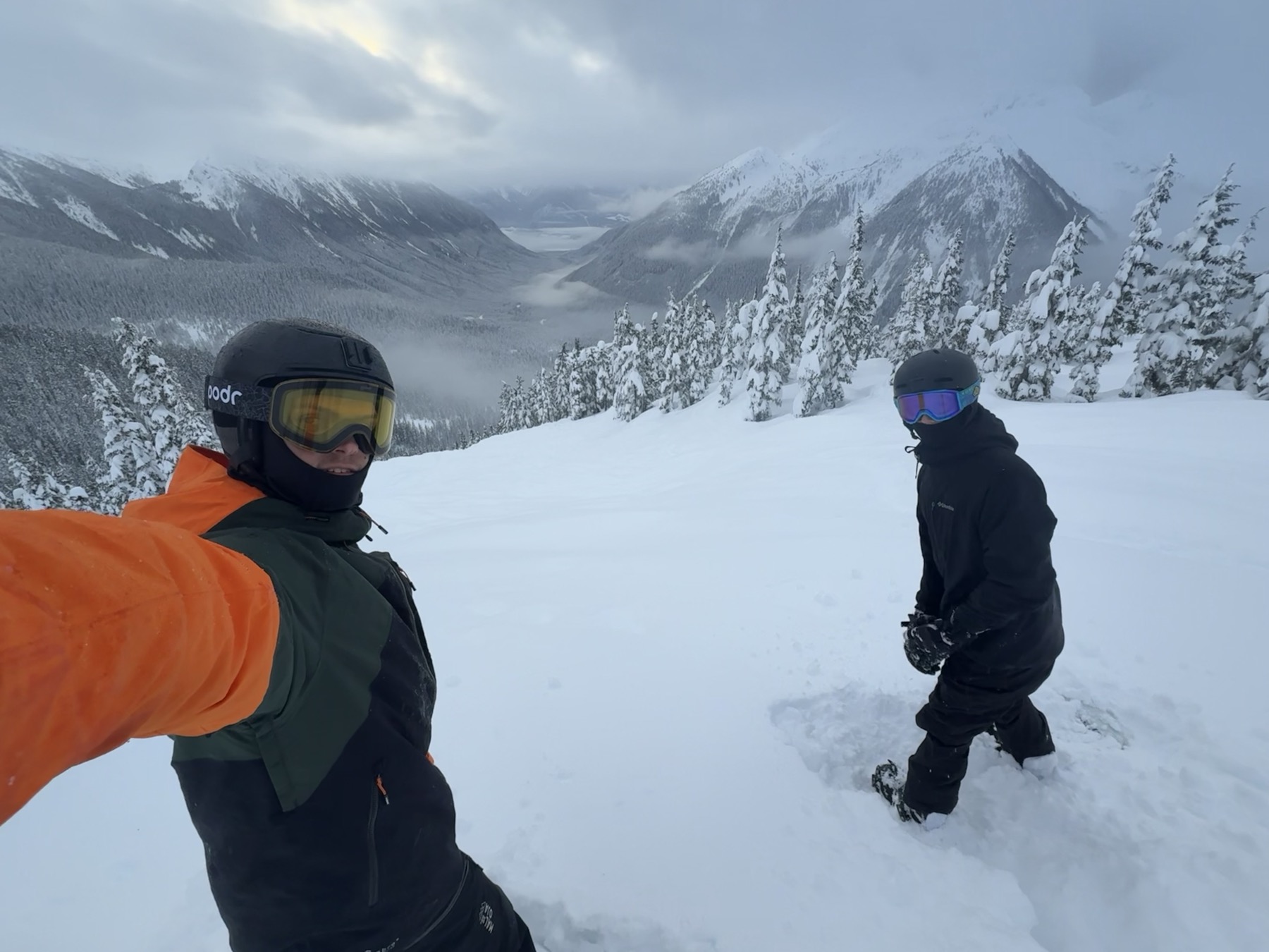 Will (left) and Calem (right) snowboarding in deep powder, with snow-covered trees and mountains behind them